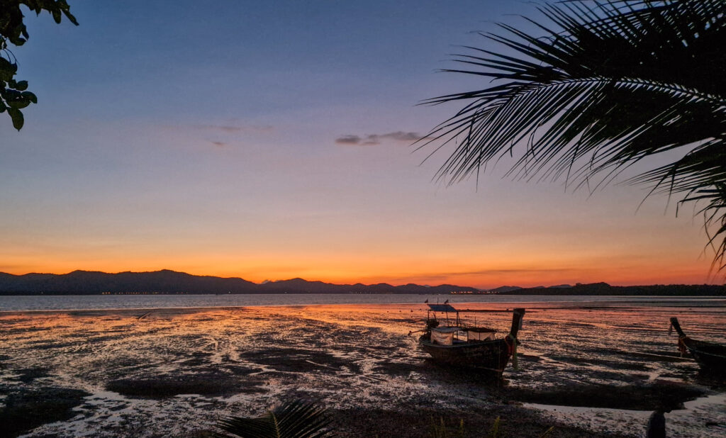 Glühender Abendhimmel über dem Meer auf Koh Yao Noi, Palme und Longtailboot im Vordergrund
